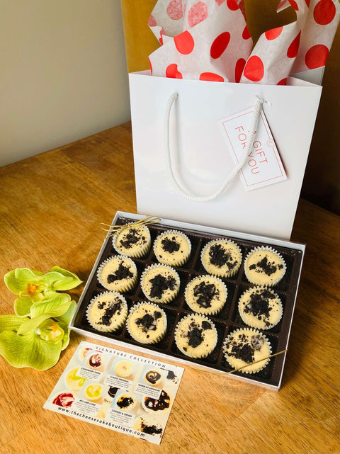 One dozen Chunky Oreo cheesecake minis in a gift box in front of a white gift bag, next to a product description tag