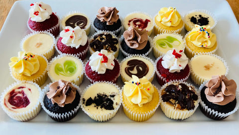 A variety of 24 mini cupcakes and cheesecakes assorted on a white platter
