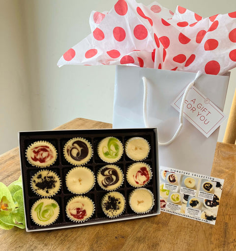 A gift box with one dozen mini cheesecakes on a table next to a gift bag and Signature collection flavor information card