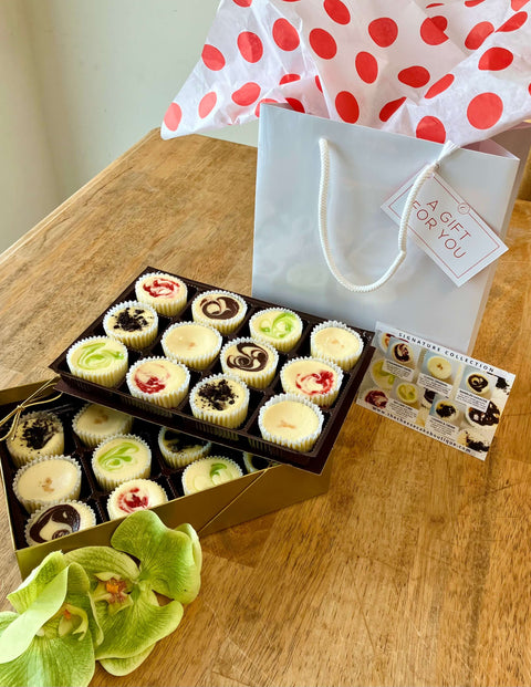 Box of assorted mini cupcakes with a gift bag and tag on a wooden surface