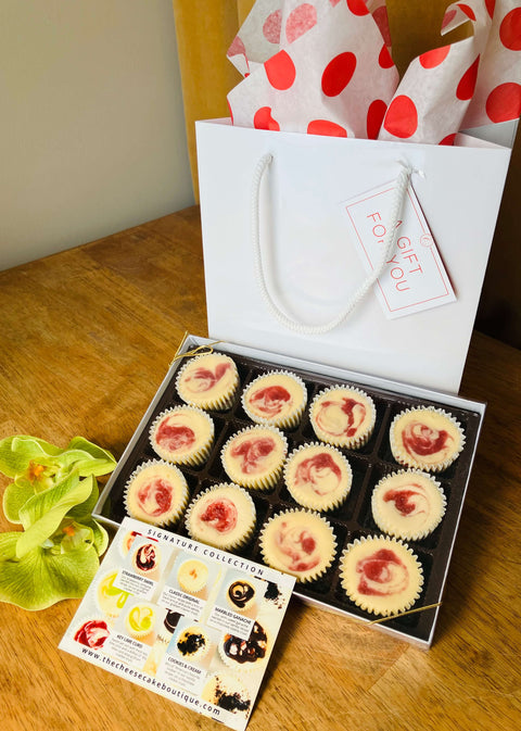 One dozen strawberry swirl cheesecake minis in a gift box in front of a white gift bag, next to a product description tag