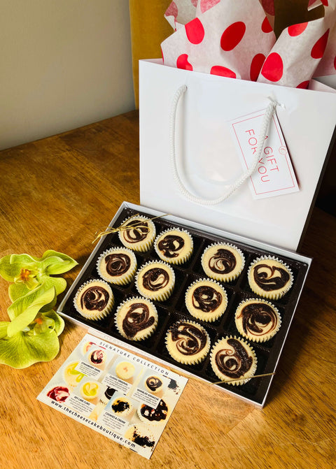 A gift box holding one dozen marbled ganache cheesecake minis in front of a white gift bag, next to a product description tag