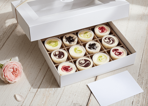 Handcrafted cheesecake minis in an open white gift box, beautifully portioned with assorted flavors including fruit swirls, chocolate, and key lime, styled with a pink rose on a light wood surface