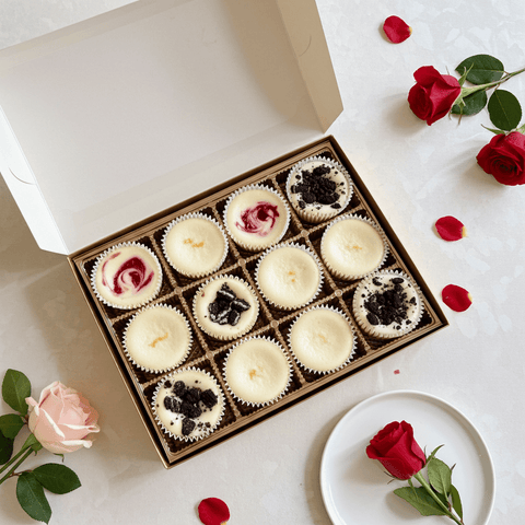 Box of mini cheesecakes with red roses and rose petals on a white surface