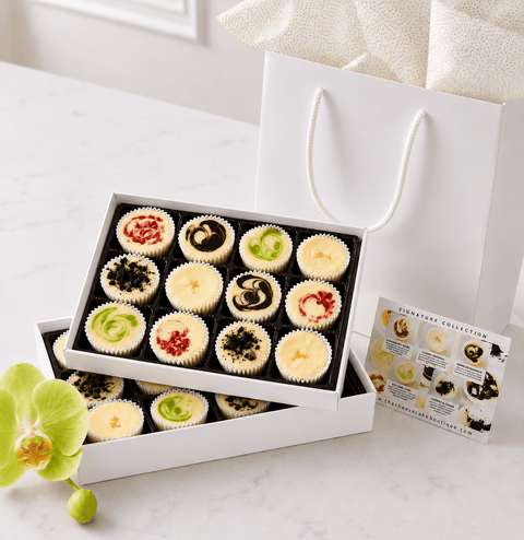 A gift box of assorted mini cheesecakes with a white gift bag and  flavor card on a light surface.