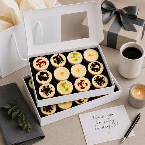A gift box of mini cheesecakes with a card, coffee, and candle on a table.