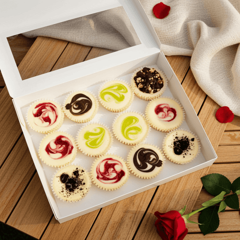 Box of assorted fruit and chocolate mini cheesecakes with colorful swirls on a wooden surface with red roses.