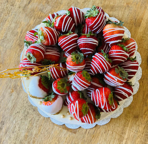 A whole strawberry cheesecake sitting on a table, topped with white chocolate-dipped strawberries, whipped cream, and a 'Proud of You' acrylic topper.