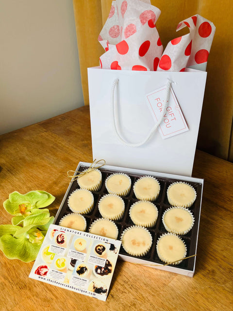 One dozen classic original cheesecake minis in a gift box in front of a white gift bag, next to a product description tag
