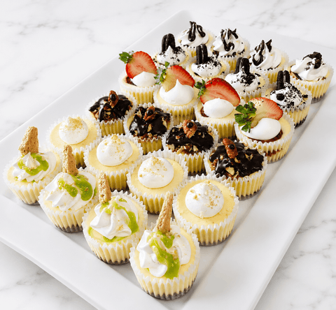 Platter of mini cheesecakes with whipped cream, strawberries, and chocolate on  white tray on a marble surface