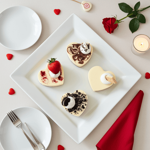 Heart-shaped individual cheesecakes on a white plate with red roses and candles, set on a beige table.