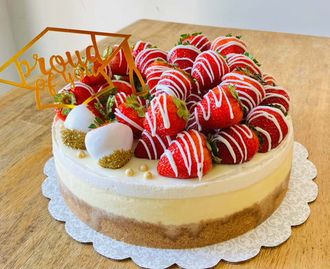 Close-up of a strawberry cheesecake on a table, showing dipped strawberries, whipped topping, and gold accents around a celebratory graduation topper.