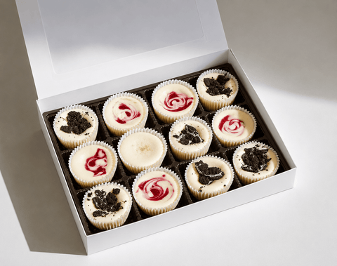 Box of twelve mini cheesecakes with fruit swirls and cookie toppings on a white background