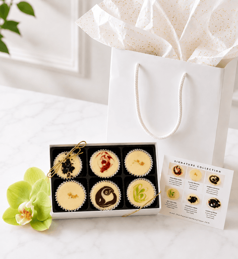 White Gift Box of six assorted mini cheesecakes with a white gift bag  on a light surface.