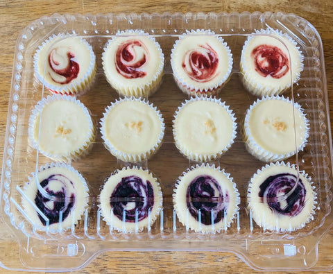 Twelve  mini cheesecakes - strawberry , blueberry and classic  in a clear plastic container on a wooden surface.