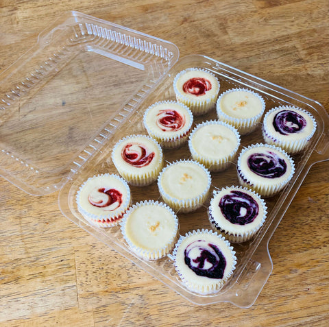 Assorted mini cheesecakes with strawberry and blueberry fruit swirls in a clear plastic container on a wooden surface