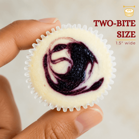 Close-up of a two-bite blueberry swirl mini cheesecake held between two fingers, showing rich blueberry topping on creamy New York-style cheesecake, 1.5 inches wide.